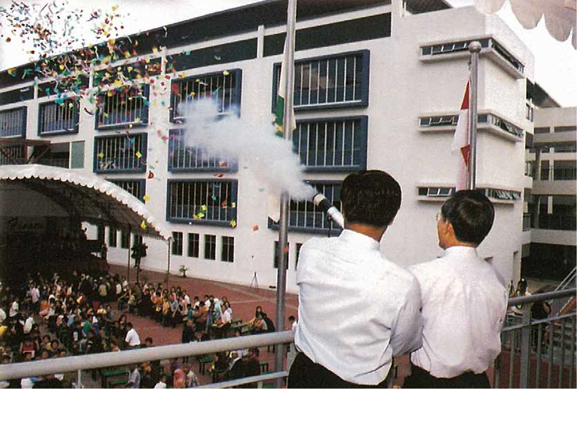 Two men in white shirts fire a confetti cannon from a balcony over a large crowd gathered in a school courtyard.